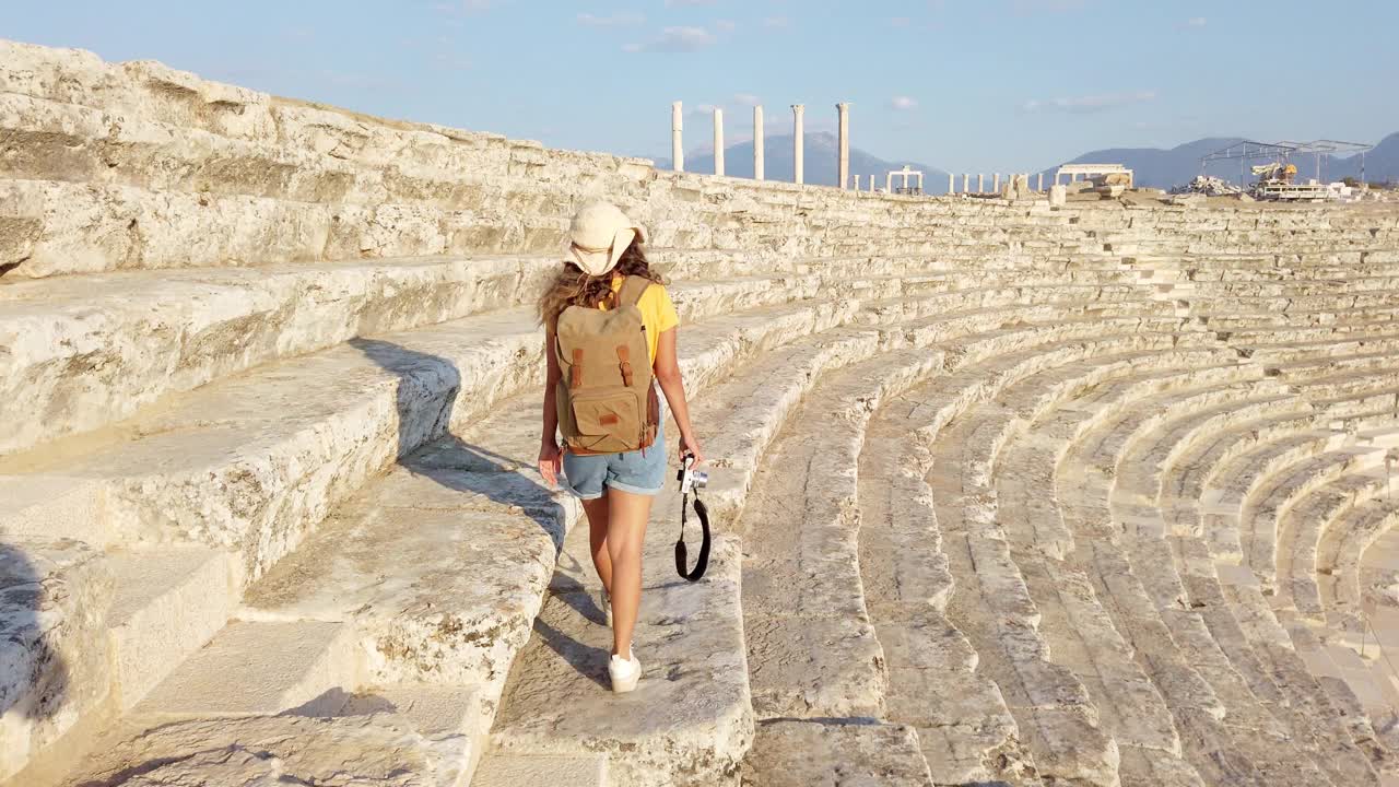 在帕姆克卡莱，日落时分，在吕卡斯老底嘉的圆形剧场的古代遗迹中，旅行者、游客摄影师女孩拿着相机走在柱子后面视频素材