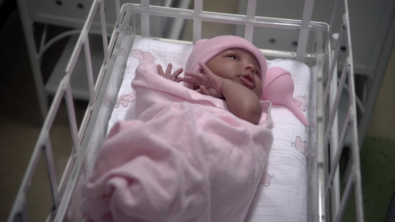 婴儿裹着粉红色的毯子，戴着粉红色的帽子，躺在医院的摇篮里。护士穿着白色制服，部分看得见检查婴儿。可能是产科病房的婴儿房。视频下载