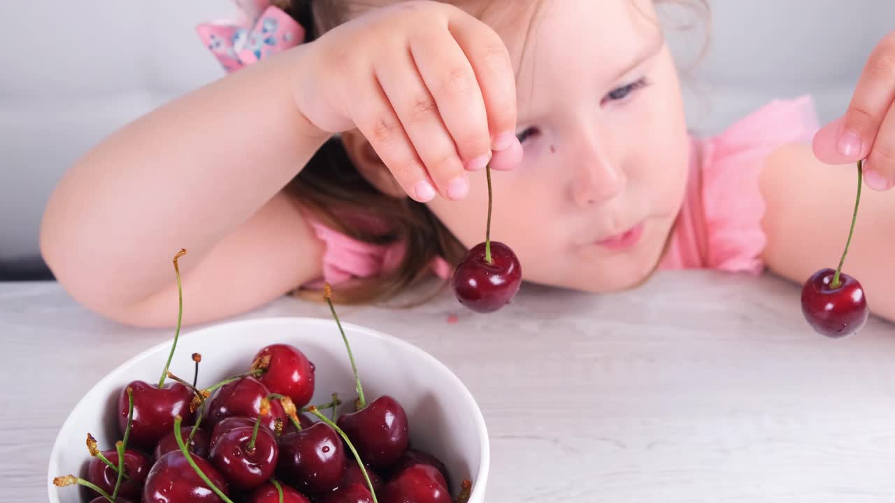 漂亮的小女孩坐在一张轻便的木桌旁，桌上放着一盘樱桃，手里拿着红色的浆果。健康饮食视频下载