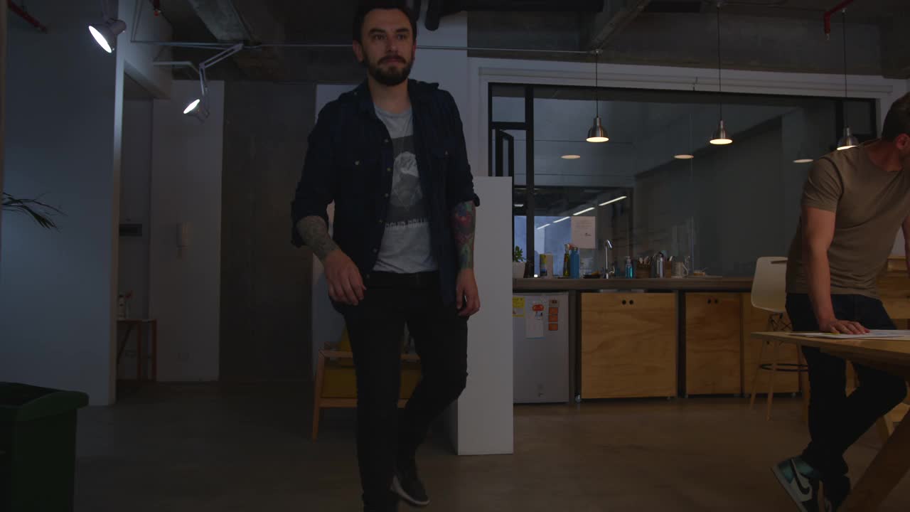 一个年轻人穿着滑板来到办公室，坐在他的办公桌前向每个人打招呼视频素材