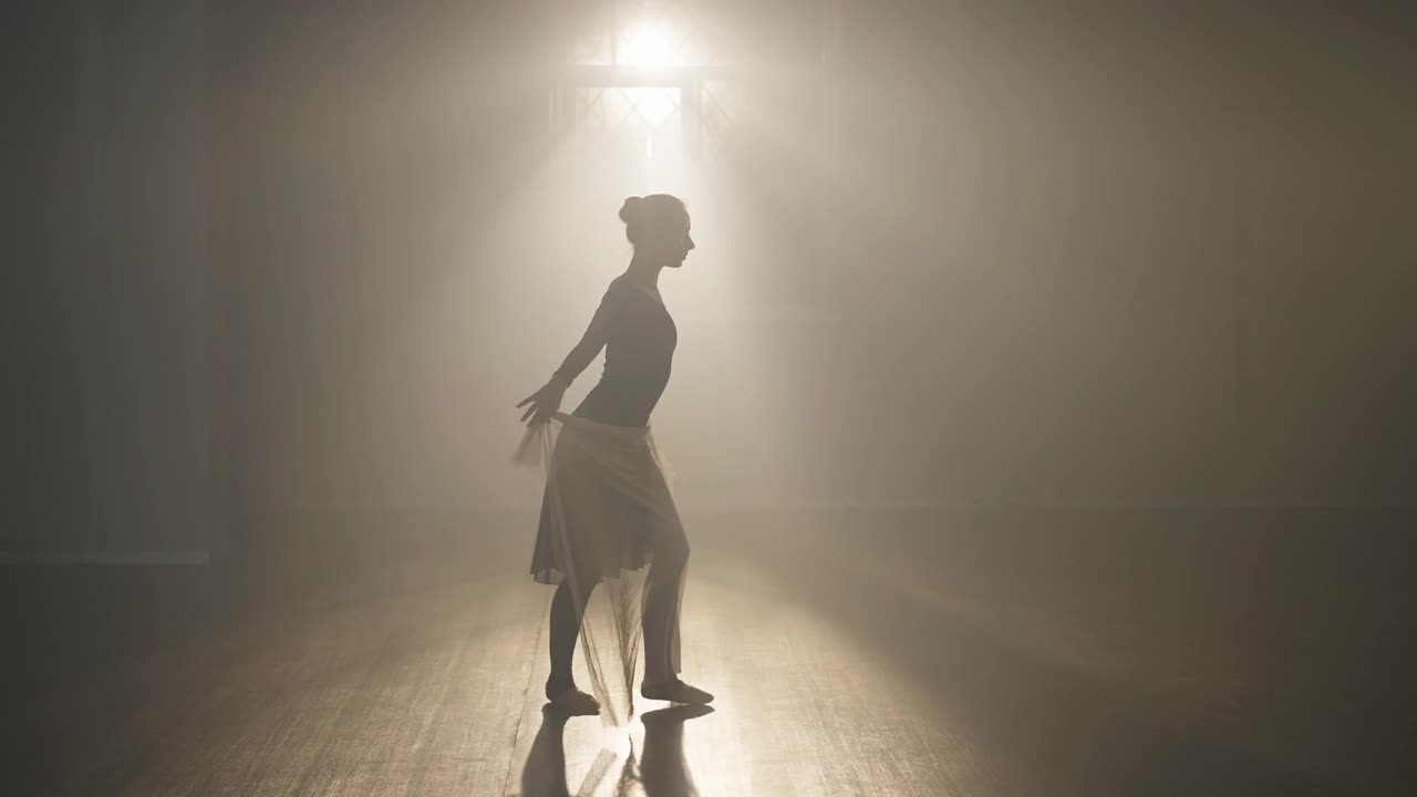 苗条的白人芭蕾舞女演员的剪影在背光的雾停止看相机。广角镜头，自信、艺术才华横溢的芭蕾舞演员在室内舞蹈室的聚光灯下排练表演。视频素材