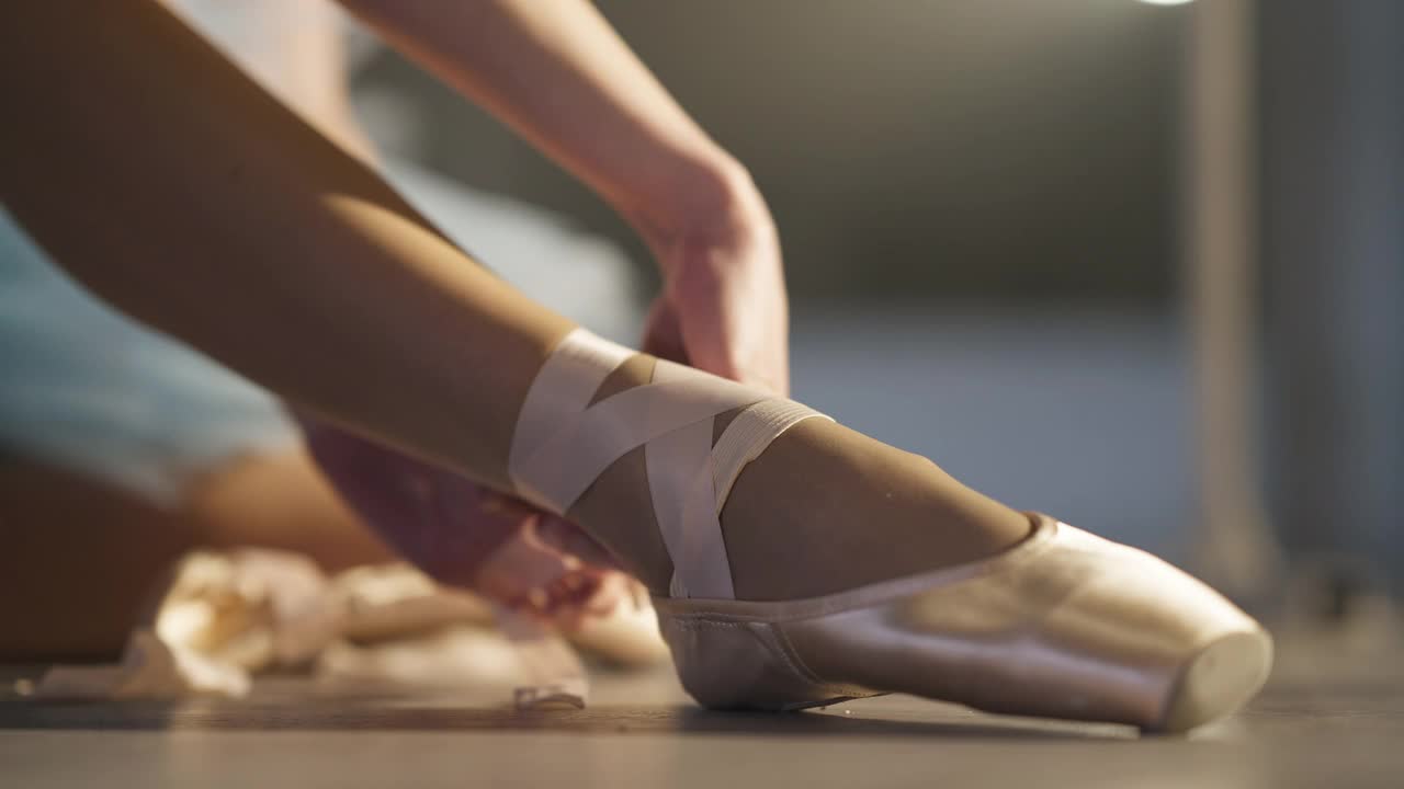 特写女性白人手系鞋带的金色脚尖鞋。在室内背光下，无法辨认的苗条优雅的芭蕾舞演员穿上了尖鞋。表演与艺术观念。视频下载