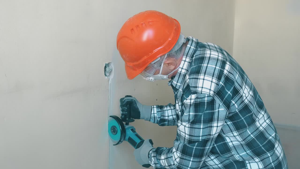 一名戴着橙色头盔和防护眼镜，带着切割金属的角磨床的老年工人在墙壁上切割，在墙壁上凿出一个凹槽，为房间里的插座铺设电线视频素材