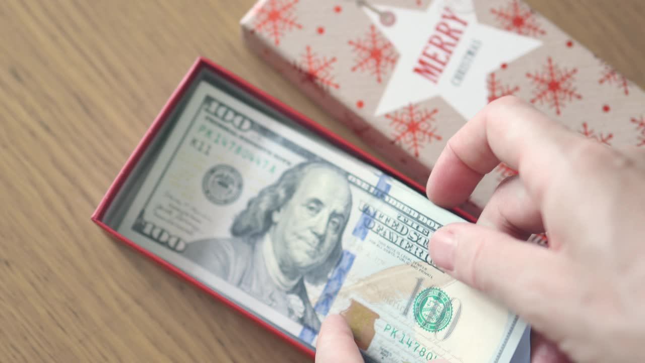 手把一百美元钞票放在礼物盒里，上面写着“圣诞快乐”。新年礼物概念视频素材