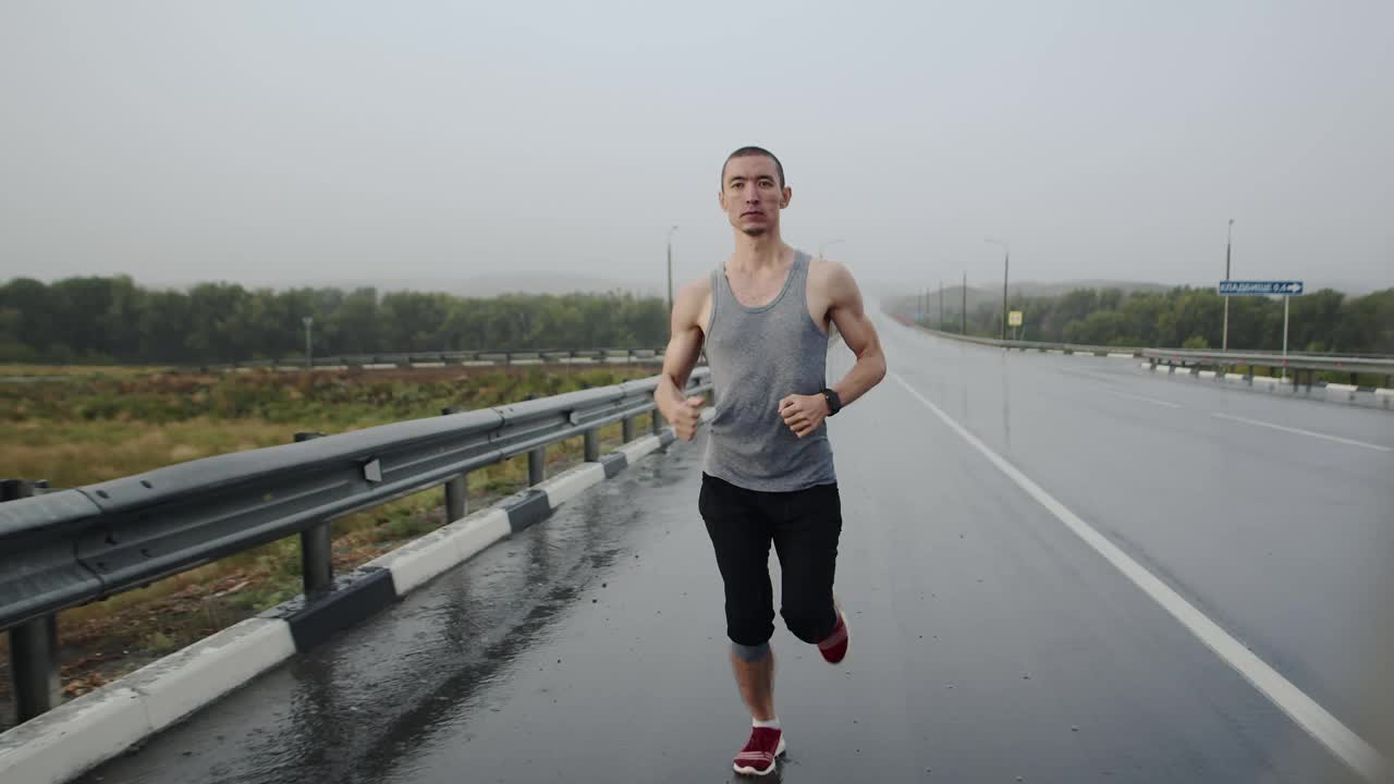 亚洲运动员与运动员身材在雨中跑在潮湿的柏油路运动鞋和触摸电子钟，前面的视图。渴望和速度的动力视频素材
