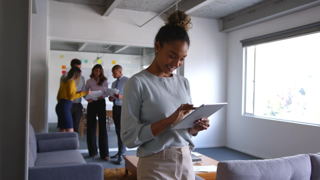 美丽开朗的黑人女性在办公室查看平板电脑上的东西，微笑着，旁边的人在后台工作视频素材