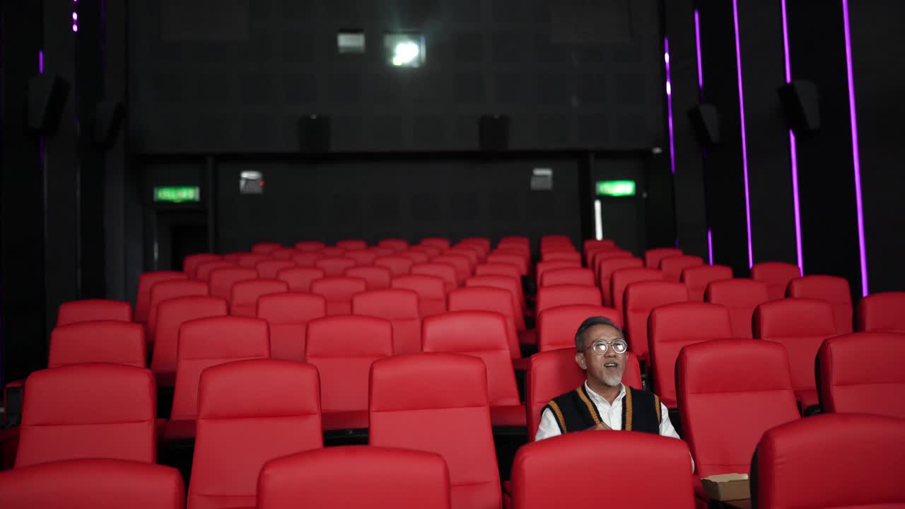 一位留着白胡子的亚裔中国男子坐在空荡荡的电影院的红色座位上一边吃一边看电影。视频下载