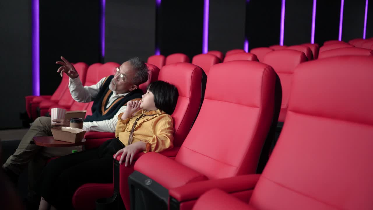 亚裔华裔爷爷和孙女坐在空荡荡的电影院的红色座位上，一边吃一边看电影视频下载