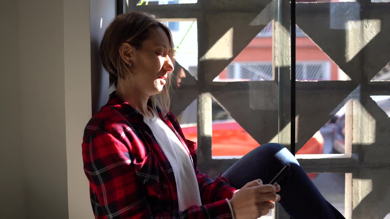 成熟的女人坐在窗台上用智能手机视频素材