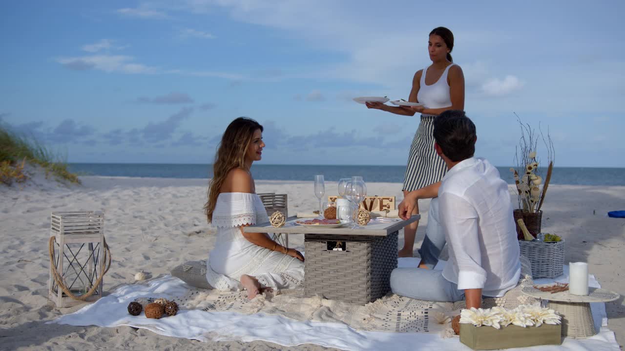 拉丁美洲的女服务员在海滩上为一对幸福的夫妇提供浪漫晚餐视频下载