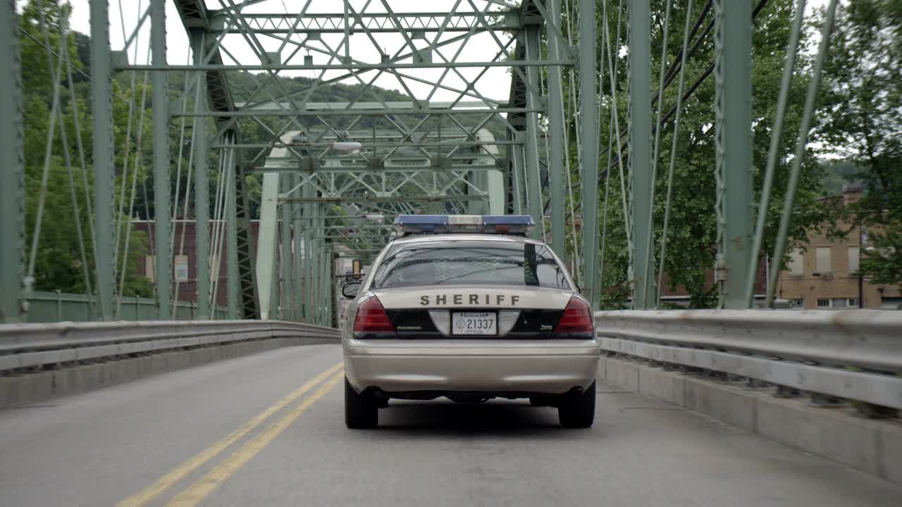 跟踪镜头显示，警长的车正开过一座桥，进入乡村地区的一个小镇。可见砖砌建筑和企业。视频素材