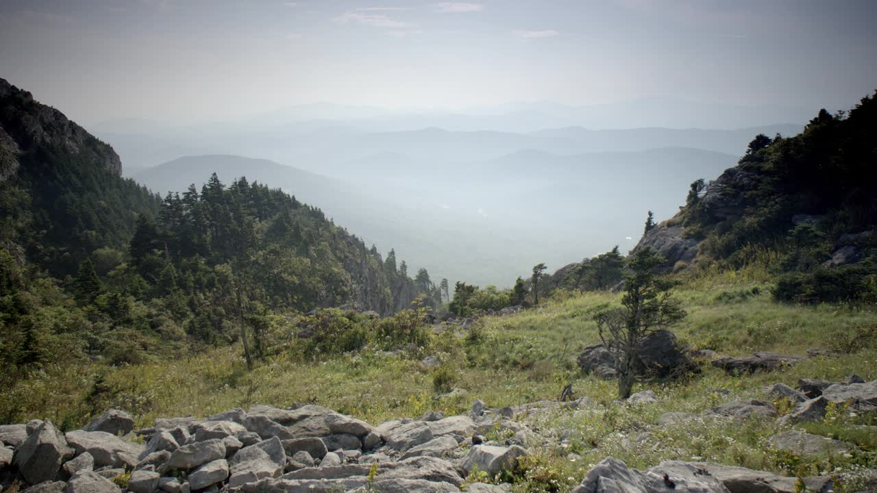 宽角度的草山坡与岩石在fg。在bg中可以看到树木覆盖的岩石悬崖和起伏的小山或山脉。朦胧的天空。视频素材