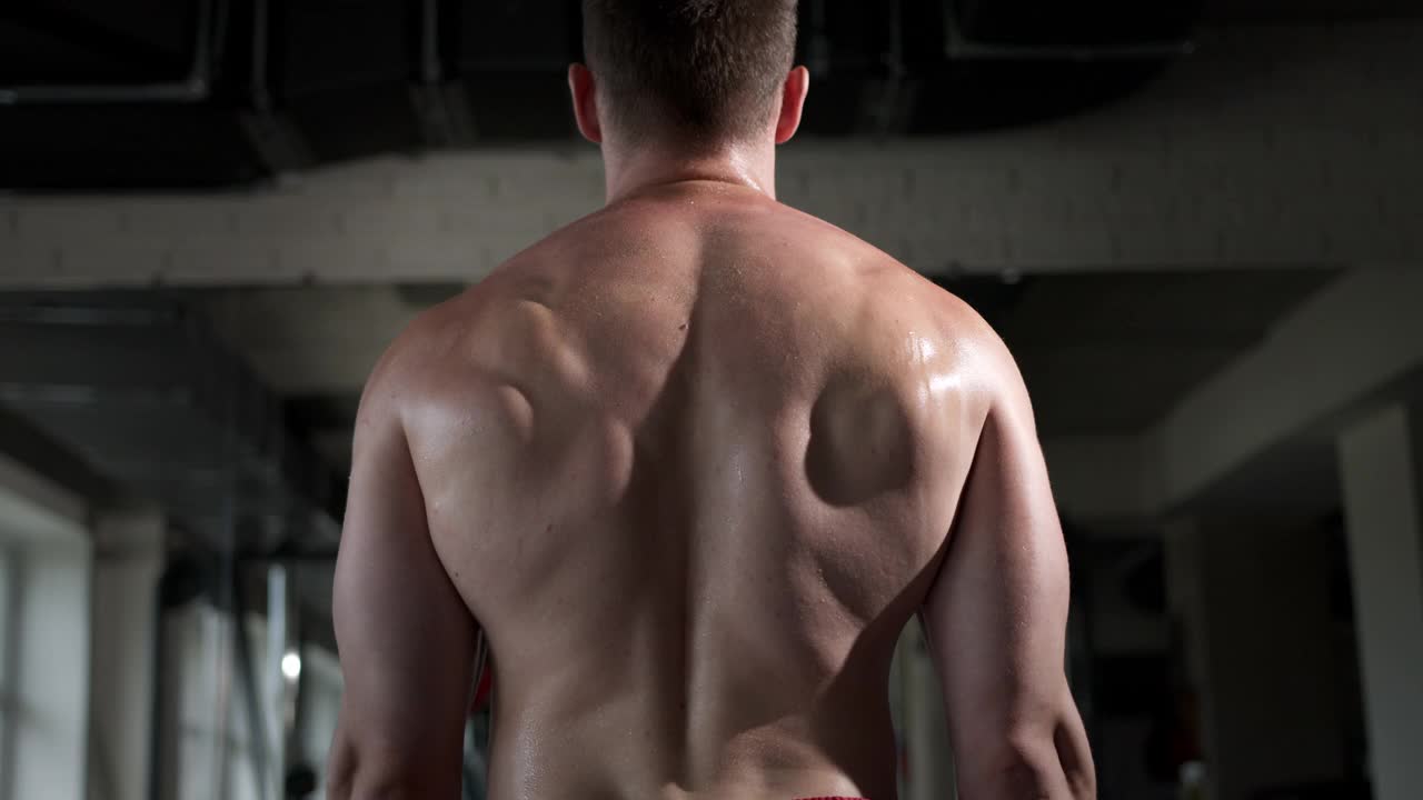 一个年轻人在健身房，站着背，练习二头肌，举起面前的杠铃。可以看到背部的肌肉是如何紧张的。视频下载