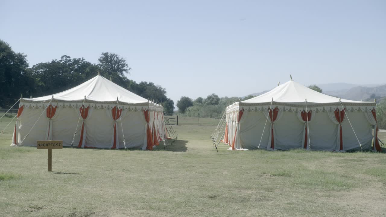 在草坪或田野上可见的两个白色和橙色马戏团帐篷的中等角度。在bg可见的树木和山脉。可以撤退。木头可见的迹象。牌子上写着“伟大的帐篷”。视频下载