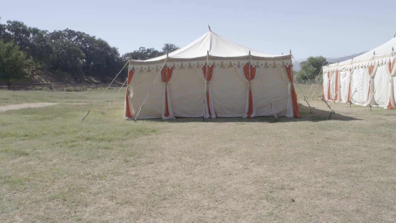 在草坪或田野上可见的两个白色和橙色马戏团帐篷的中等角度。树木和山脉部分可见在bg。镜头向下平移到写着“冥想”的木质标志。可以撤退。视频下载