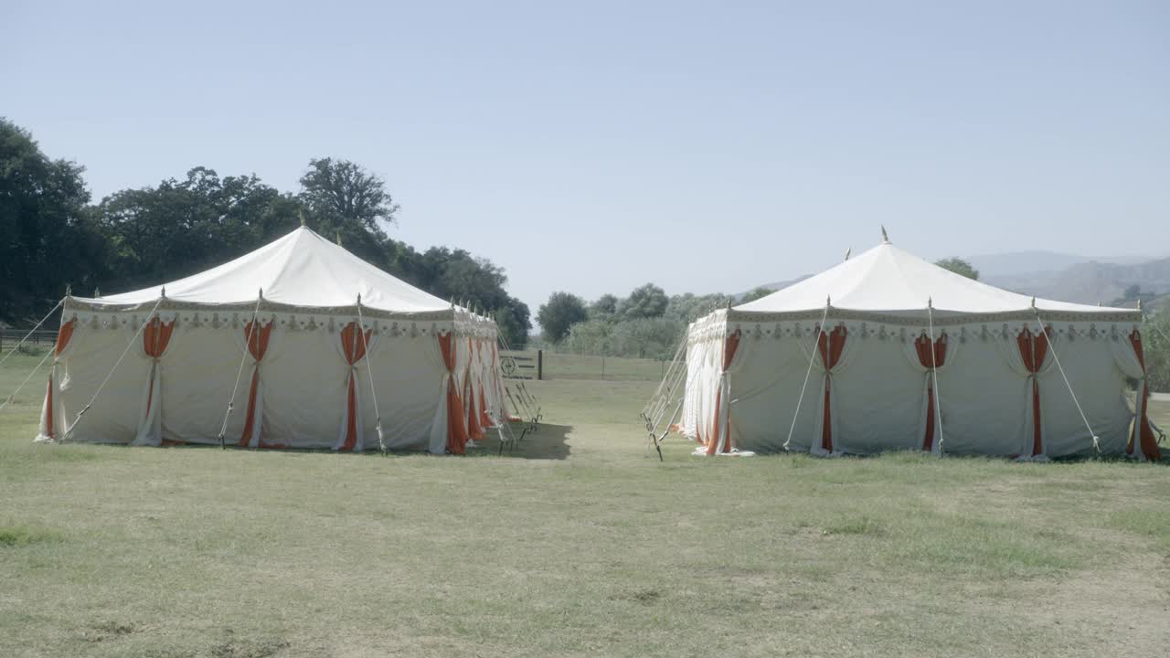 在草坪或田野上可见的两个白色和橙色马戏团帐篷的中等角度。在bg可见的树木和山脉。可以撤退。视频下载