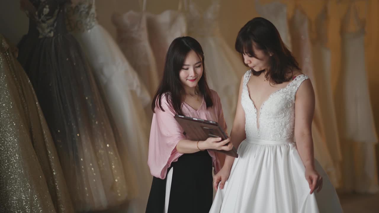 亚洲的中国女新娘商店老板向她的顾客解释新娘使用数码平板选择套餐定价定制设计的婚纱视频下载