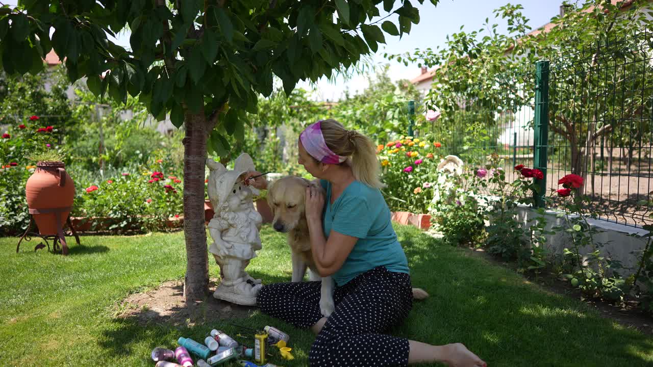 女艺术家在她的艺术花园侏儒雕像上工作，在她的花园与金毛猎犬户外绘画。视频下载