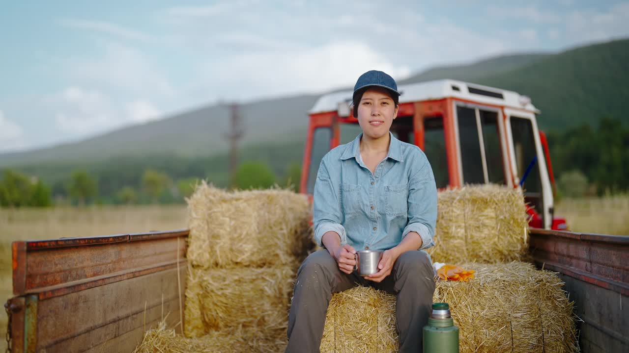 年轻的女农民坐在干草堆上的拖拉机拖车上，在农田上休息视频素材