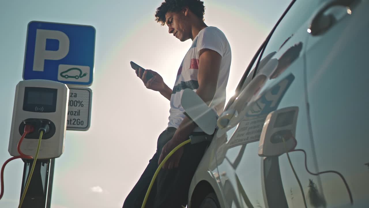 一名年轻人在停车场给汽车充电时使用智能手机视频下载