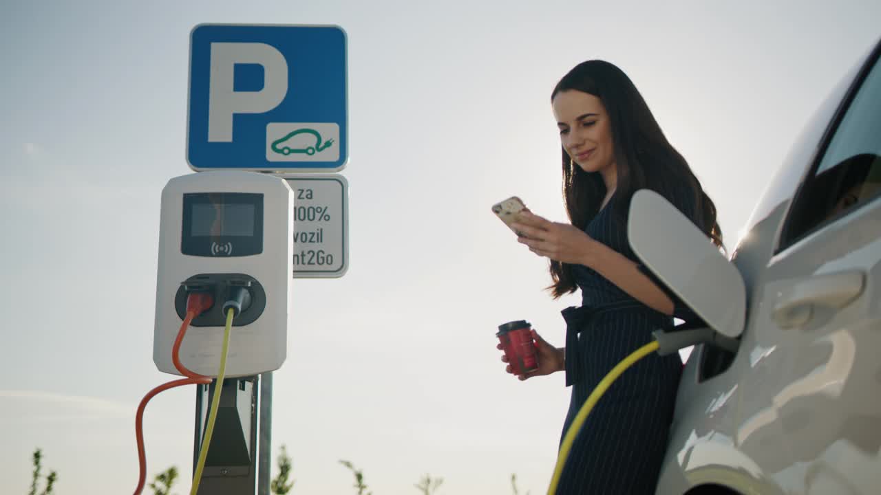 一位年轻女子在充电的汽车旁用智能手机喝咖啡视频下载