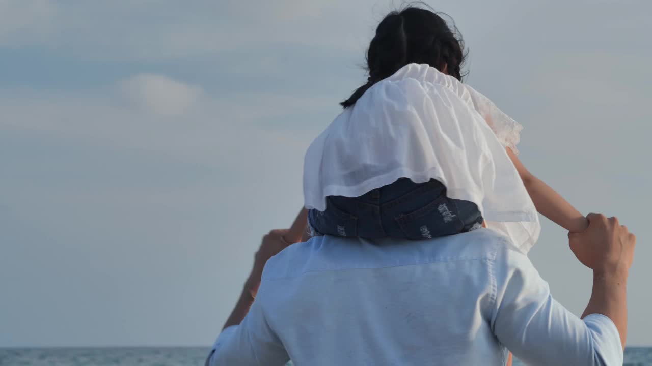后视图，亚洲家庭的父亲和4岁的女儿骑在父亲的脖子上，牵着她的手，一起在户外的海滩上看风景。幸福的家庭假期。和人民的关系。张开双臂的概念。视频素材