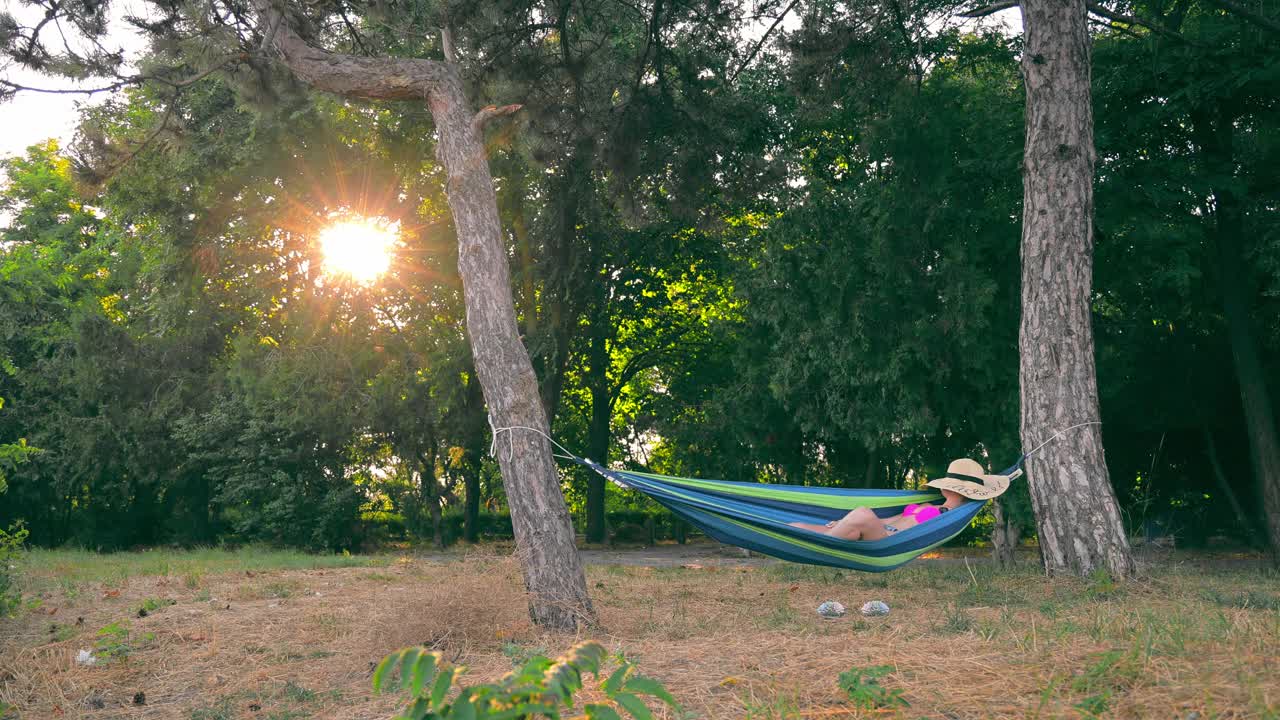 一个快乐的年轻女人躺在公园的吊床在日落和床上帽子在她的脸。在公园的吊床上好好休息一下。日落时分，一个穿着泳衣的处女躺在吊床上视频素材