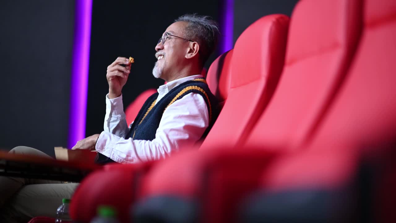侧视亚洲华人活跃的老人享受他的零食爆米花，同时在电影院看电影，独自一人视频下载