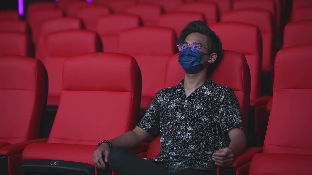 亚洲华人少年戴着口罩在电影院看电影，呆若木鸡，跷着二郎腿视频下载