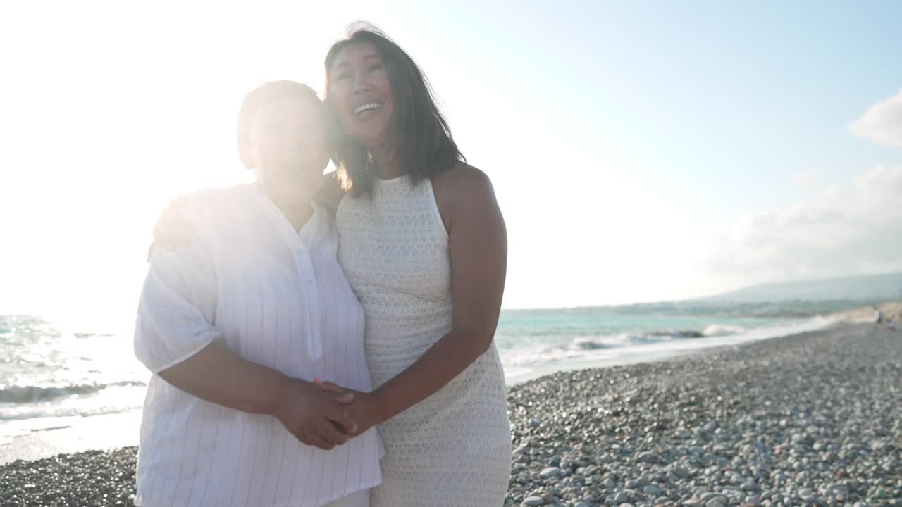 美丽的成年亚洲女儿和开朗的年长母亲站在地中海海滩上，在阳光下跳舞唱歌。幸福的女人在绿松石海岸度假的肖像。视频下载