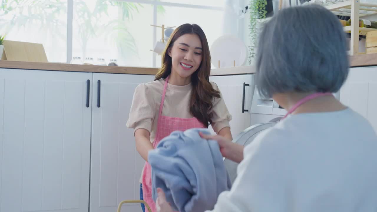 亚洲可爱的家庭做家务和家务在家里的厨房。年轻的女儿帮助和支持年长的母亲把衣服放进洗衣机里洗。客房清洁概念视频素材