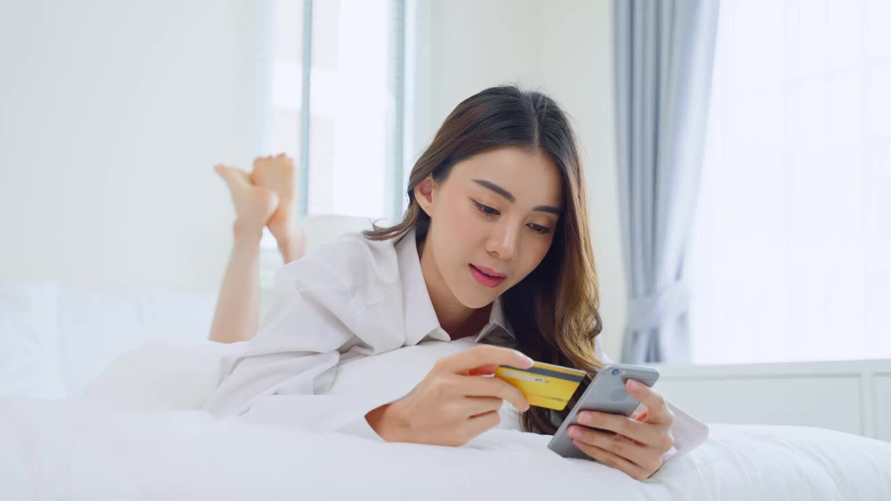 亚洲美女喜欢在床上购物在家里早上。偶然的女性躺在卧室里，在家里的网店使用手机购物后，拿着信用卡进行网上银行支付视频素材