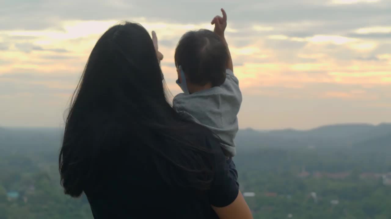 夕阳下的幸福家庭背面，亚洲母亲和17个月大的男婴戴着防护口罩，在山上自由地欣赏大自然，教孩子可持续发展，在大自然中快乐。视频素材