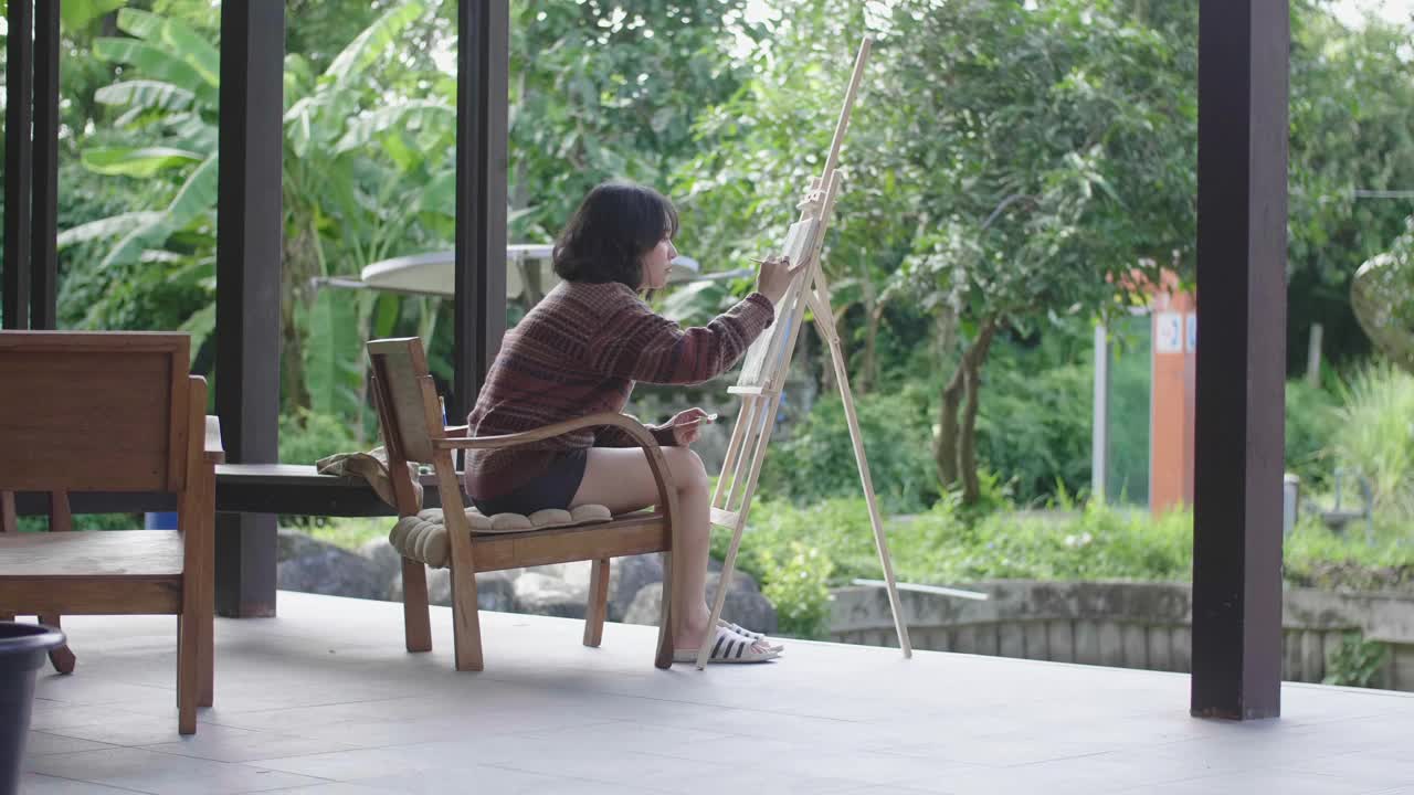 年轻的亚洲女画家在后院用画笔和画架创作艺术品。视频素材