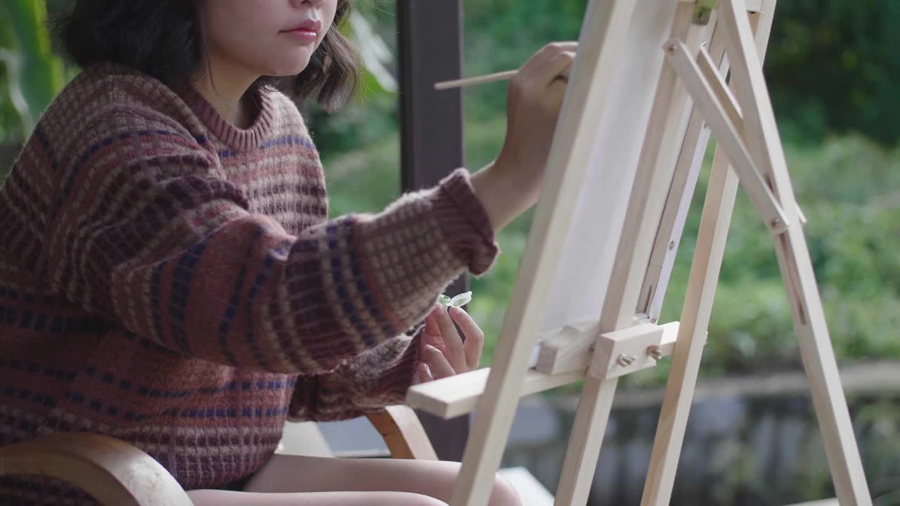 年轻的亚洲女画家在后院用画笔和画架创作艺术品。视频素材