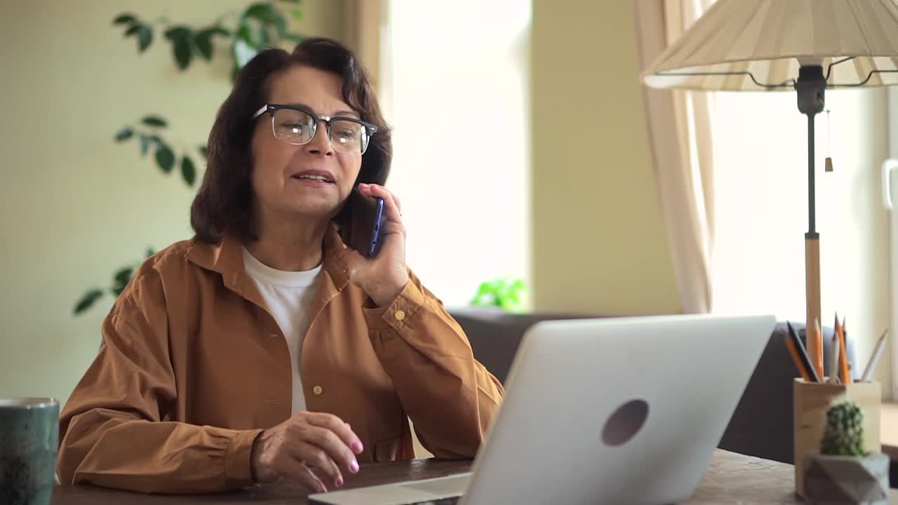 女人在家里办公室的桌子上讲电话和用笔记本电脑工作视频素材