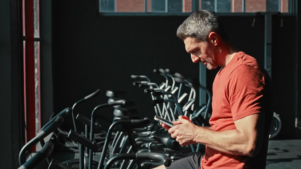 一个成年男子一边使用健身器材一边用手机锻炼的4k视频片段视频素材