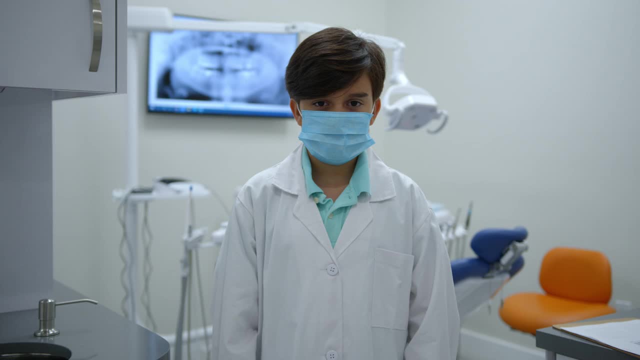可爱的小男孩在一个牙医练习玩假装穿着实验服和保护面罩交叉手臂和面对摄像机视频下载