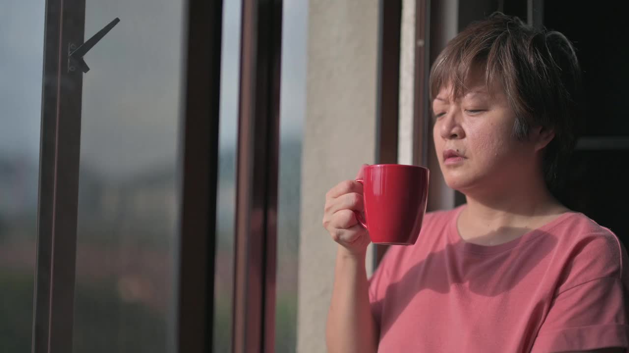 慢动作亚洲华人中年妇女享受她的热饮看着窗外放松的早晨视频下载