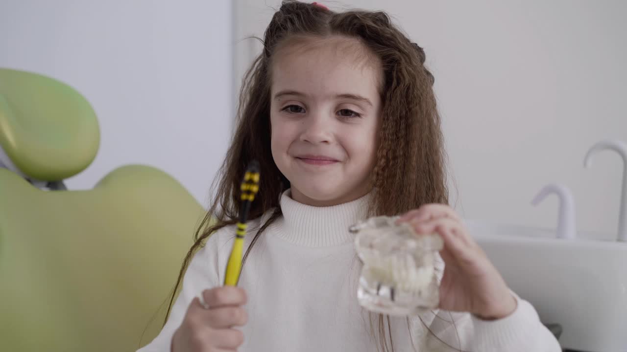 可爱的女孩拿着牙刷，下巴布局，看着牙医椅，对着镜头微笑视频素材