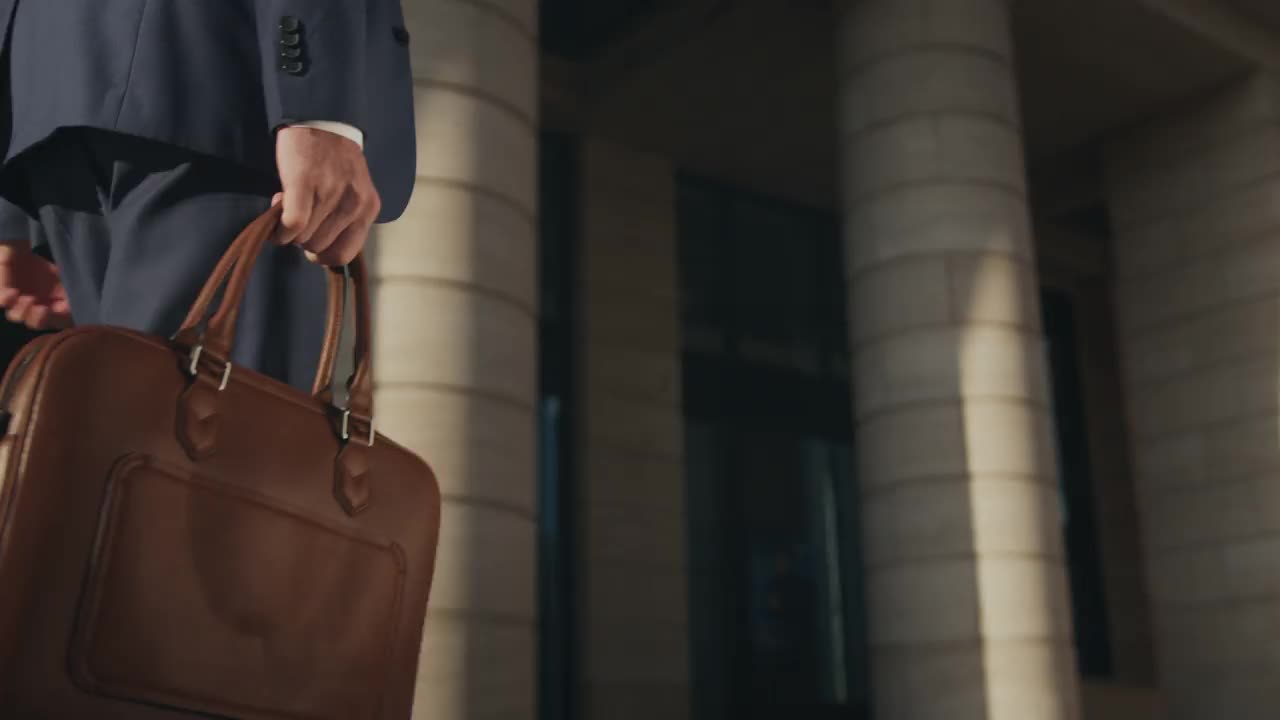 一个穿着深蓝色西装的男人走着，右手拿着棕色皮革笔记本电脑包，这是焦点，只能看到身体的一部分视频下载