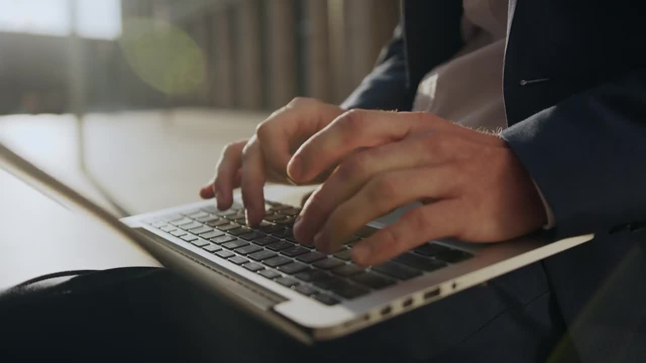 年轻商人的双手，在笔记本电脑上打印，躺在膝上，在夕阳的照耀下，对着现代建筑的立面视频下载