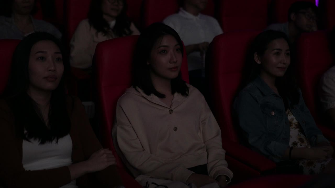 一位亚裔中国年轻女子在电影院看电影时使用智能手机。打扰她周围的观众视频下载