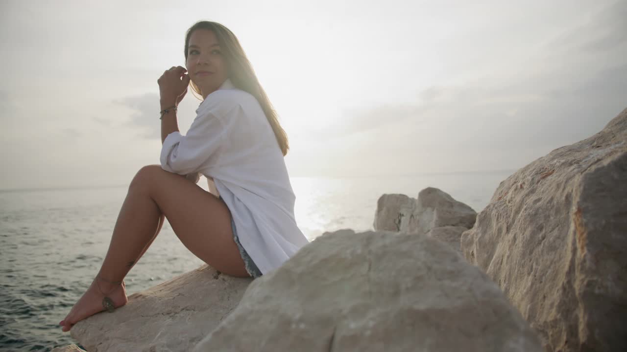 一个快乐的年轻女子坐在沙滩上的岩石上视频素材