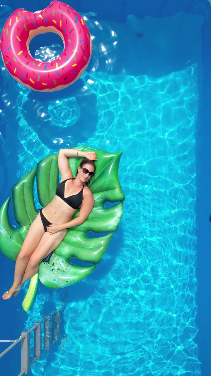 垂直:年轻的白人妇女躺在她的家游泳池的一个大叶子浮子。视频下载