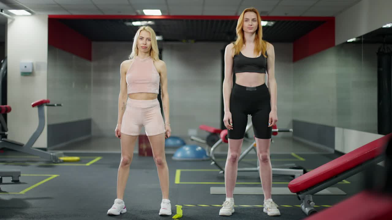 两名自信的年轻白人女性在健身房伸展双手训练的广角照片。红发和金发女士在室内做热身运动的正面照片。健康的生活方式和运动理念视频素材
