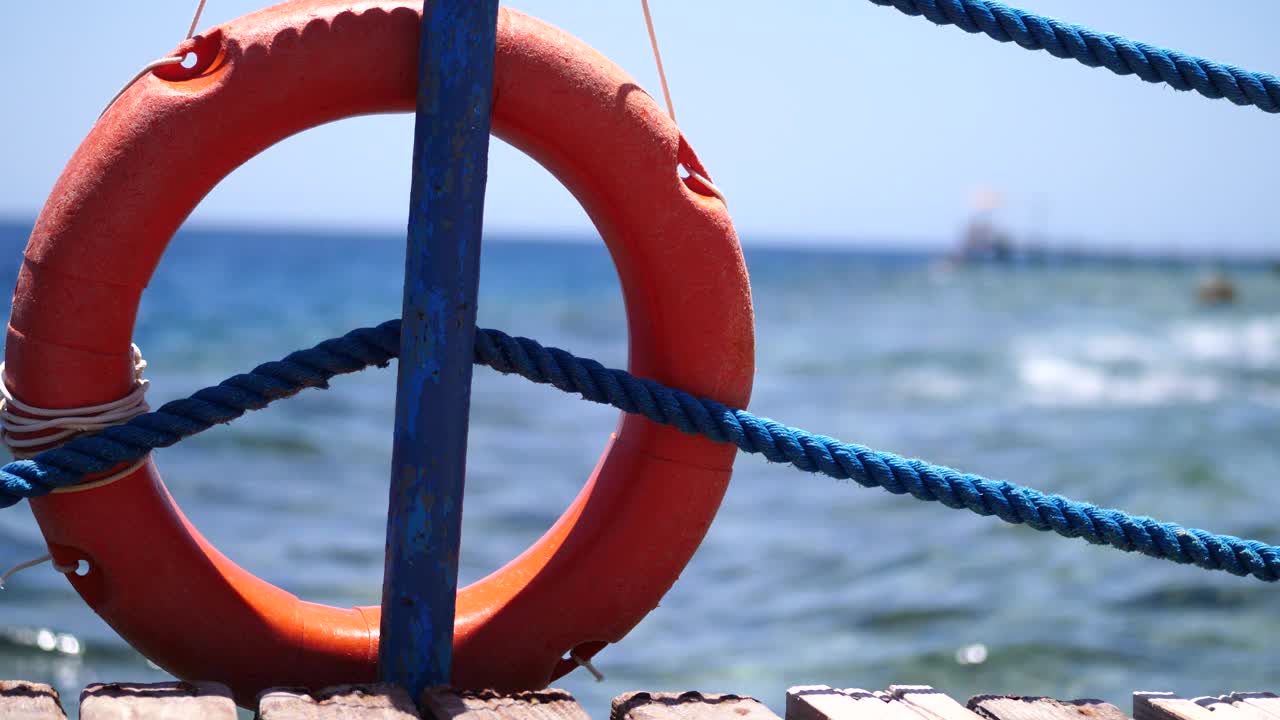 橙色的环形救生圈悬挂在码头上的杆子上，背景是蓝天和在大海中游泳的人视频素材
