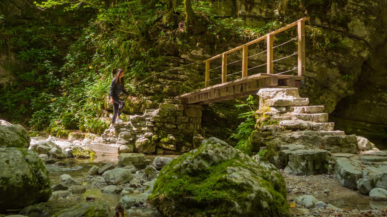 健美的女子在大自然中行走，穿过一座桥，在雄伟的绿色峡谷中流过溪水视频素材