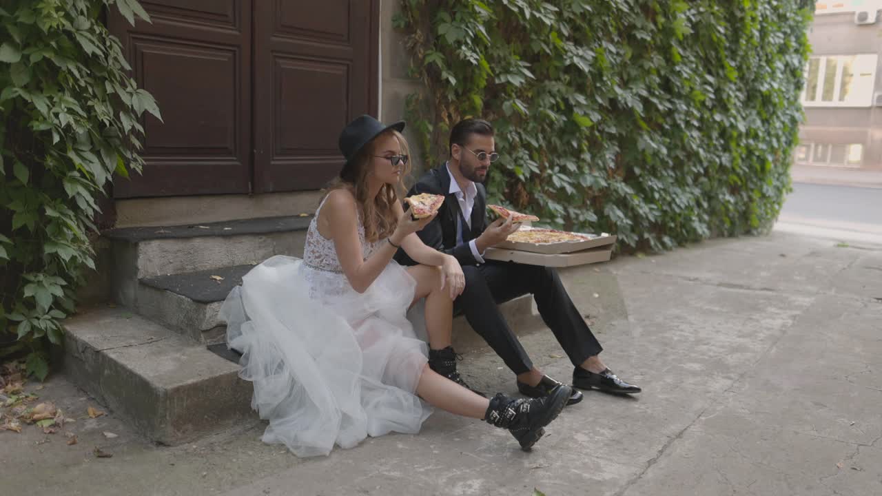 新娘和新郎坐在小巷的楼梯上，一边吃着披萨一边微笑视频下载