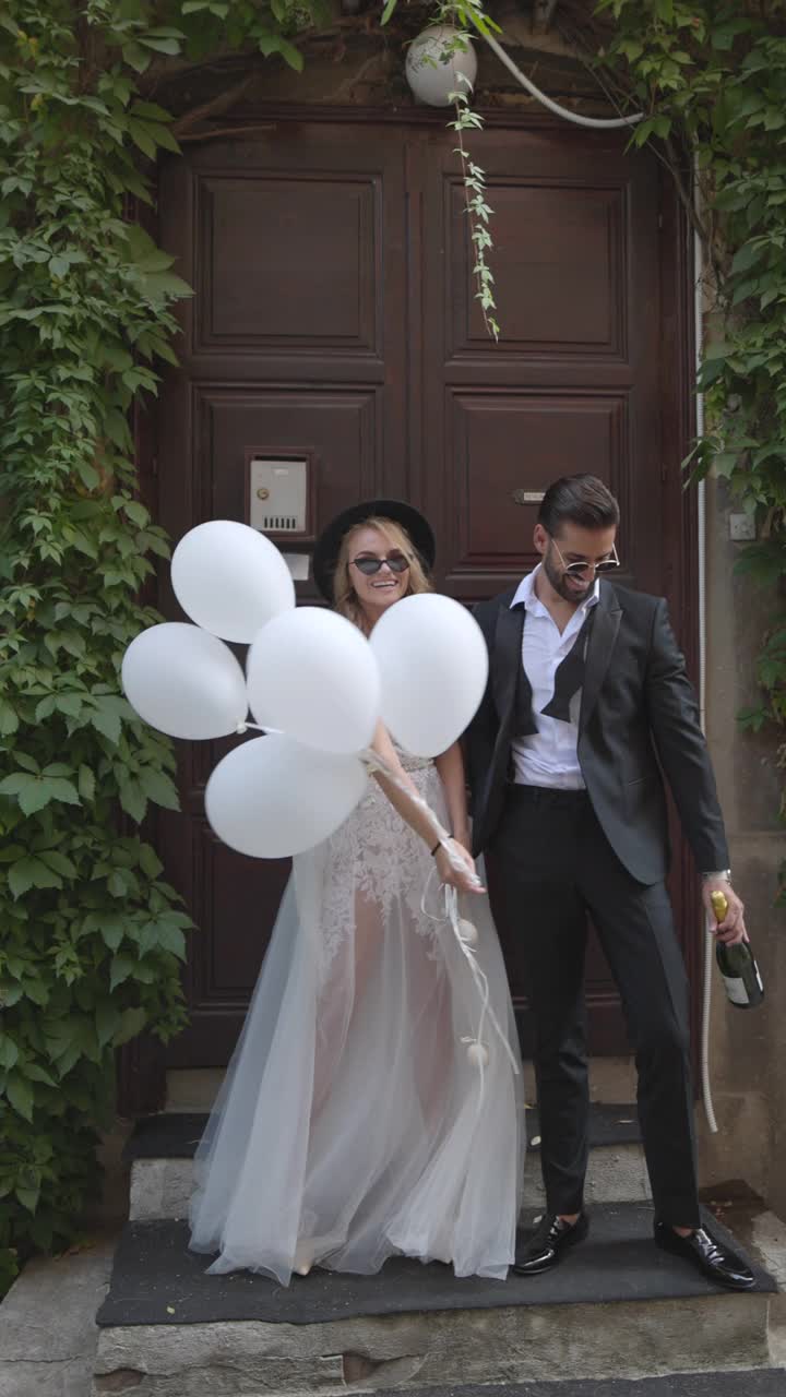 微笑的新郎和新娘站在乡村的门前，手里拿着气球和香槟视频下载